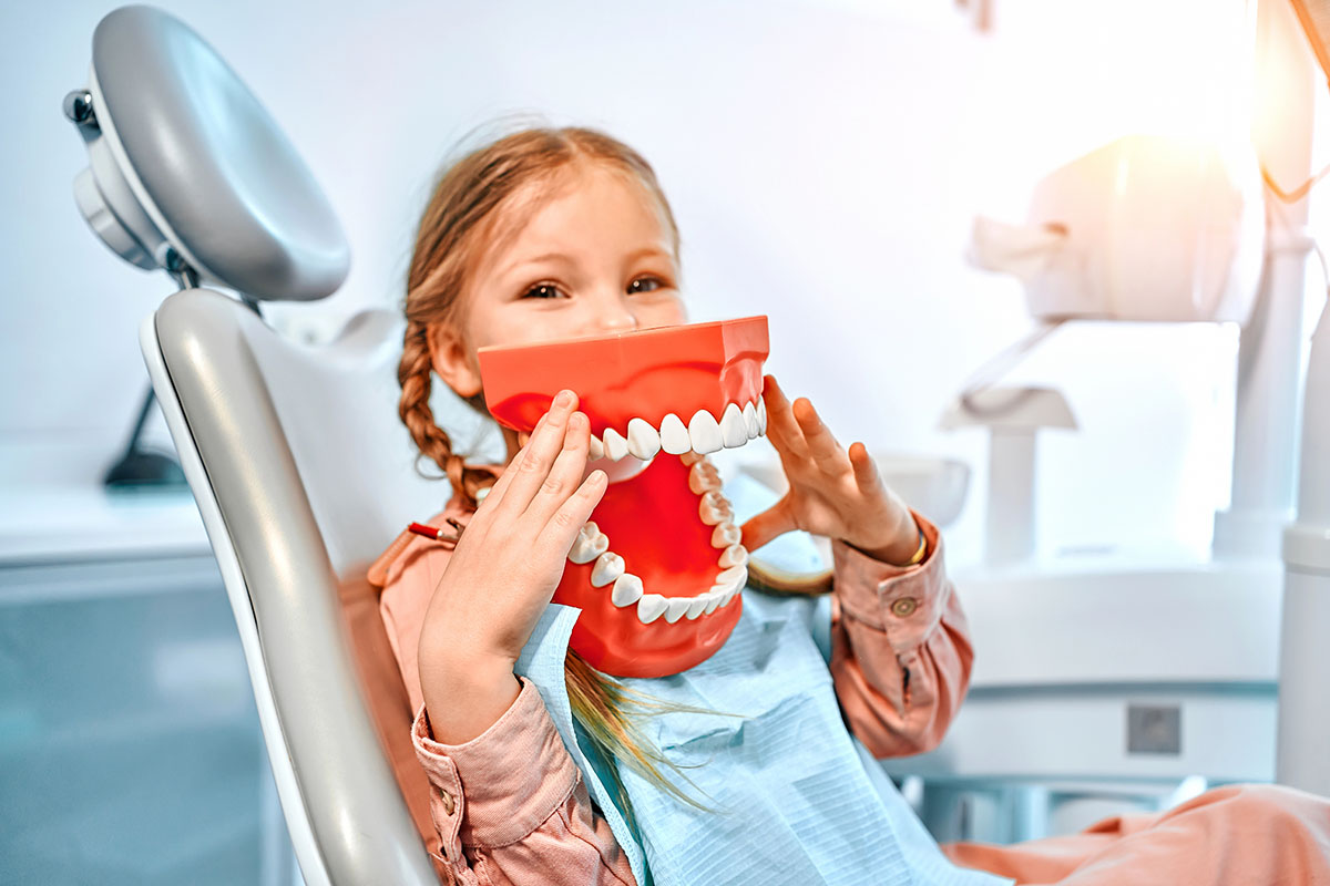 A smiling girl holds a large, exaggerated model of teeth in front of her face while sitting in a dental chair, wearing a dental bib.