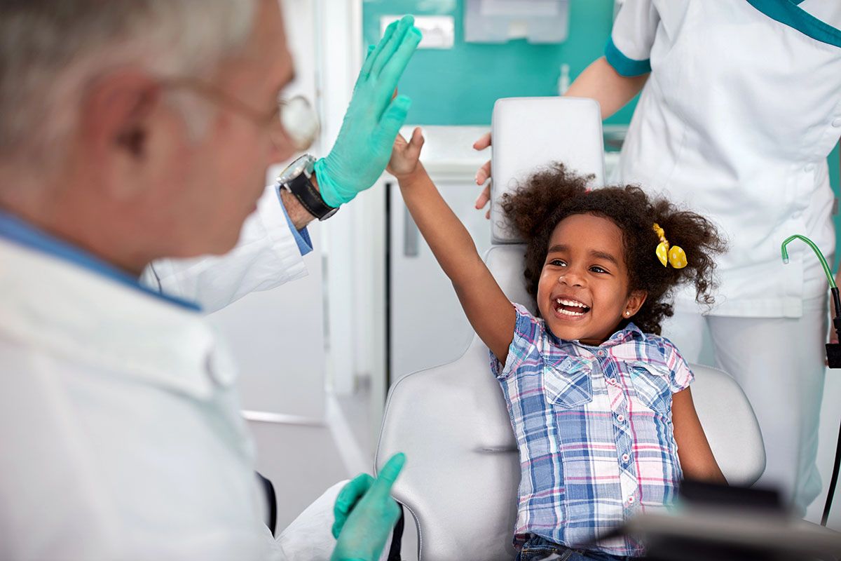 A young girl sitting in a dental chair is high-fiving an older male dentist.