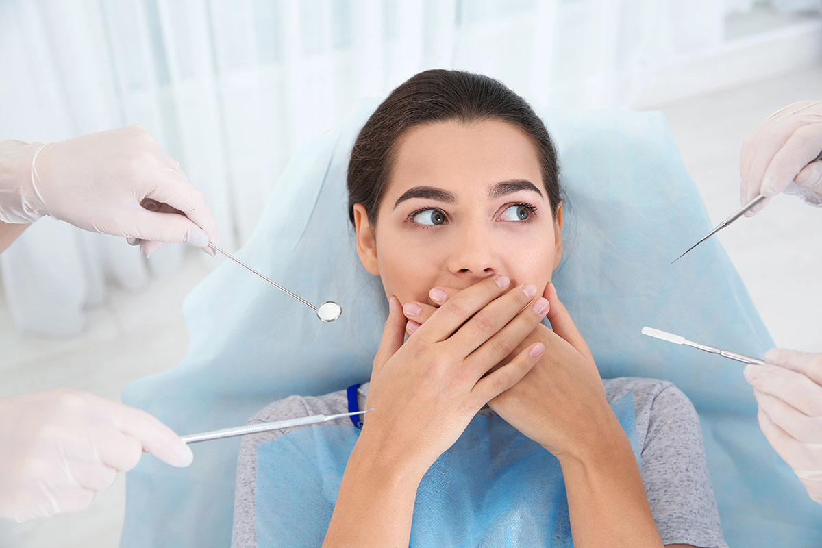 A woman looking anxious in a dental chair holds her hands over her mouth while dental instruments are positioned near her by gloved hands.