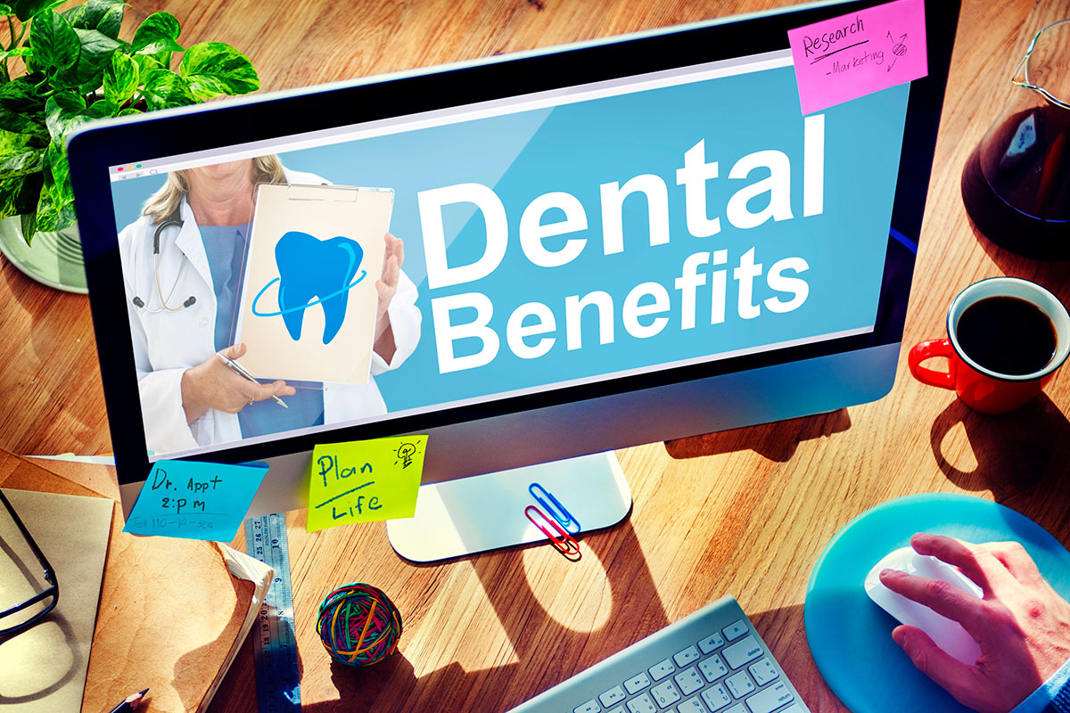 A top-down view of an office workspace featuring a desktop monitor displaying "Dental Benefits" with a tooth graphic, surrounded by sticky notes, a keyboard, mouse, and coffee mug.