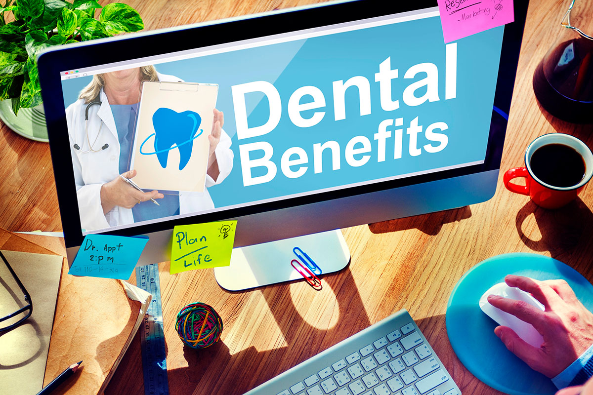 A computer monitor displays the text "Dental Benefits" with a blue tooth graphic, alongside a hand holding a clipboard. The workspace features sticky notes, a coffee mug, a ball of colorful rubber bands, and a keyboard.