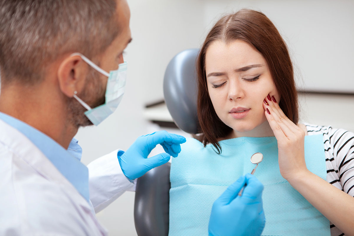 A female patient holding her cheek in pain while sitting in a dentist's chair as a dentist prepares to examine her.