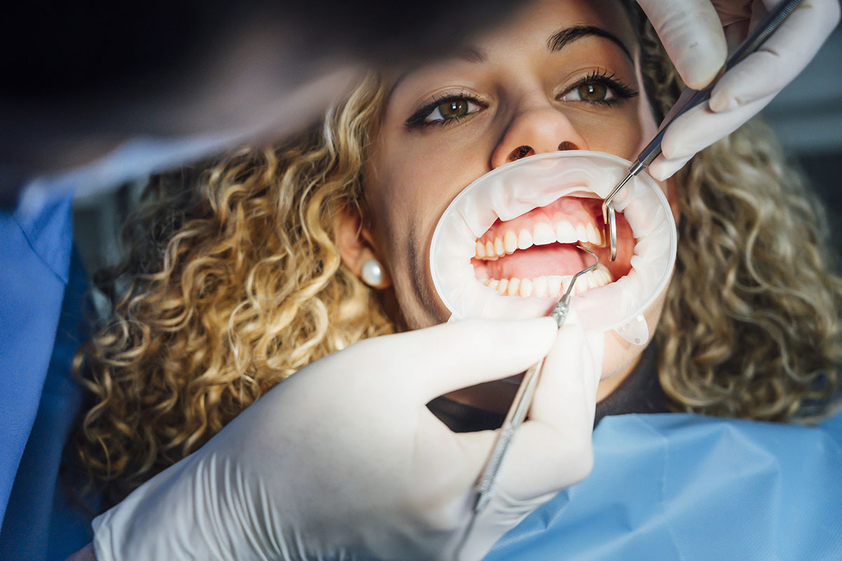 A patient in a dental chair has her mouth open with a dental bib on, while a dentist examines her teeth using dental tools, wearing gloves.