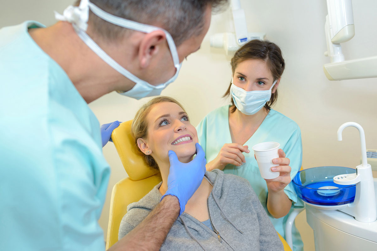 A woman is sitting in a dental examination chair while a dentist and his assistant inspect her teeth in a well-lit dental office.