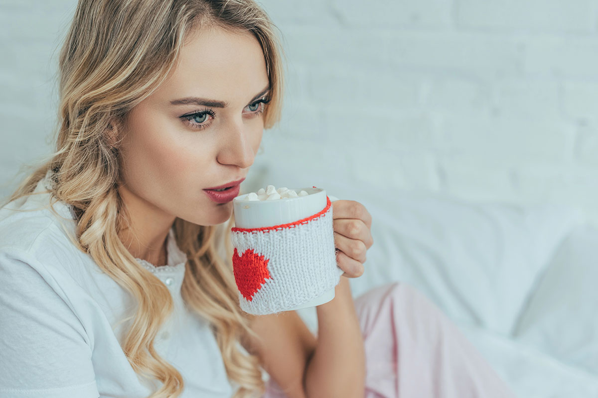 A woman with long, wavy blonde hair holds a white mug with a knitted cozy featuring a red heart, taking a sip of hot chocolate topped with marshmallows. She wears a white shirt and sits on a bed with light-colored bedding, in front of a white brick wall.