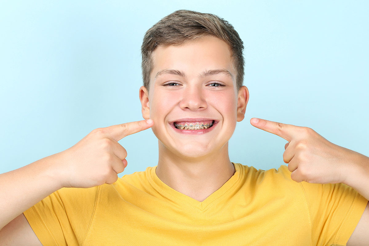 A young man with brown hair and a yellow shirt is smiling while pointing at his dental braces, against a blue background.