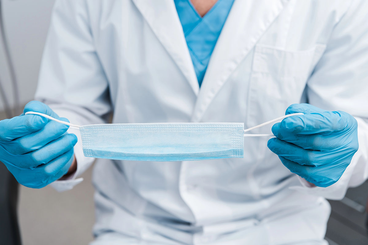 A healthcare professional in a white lab coat and blue gloves holds a light blue surgical mask by its ear loops, preparing to wear it.