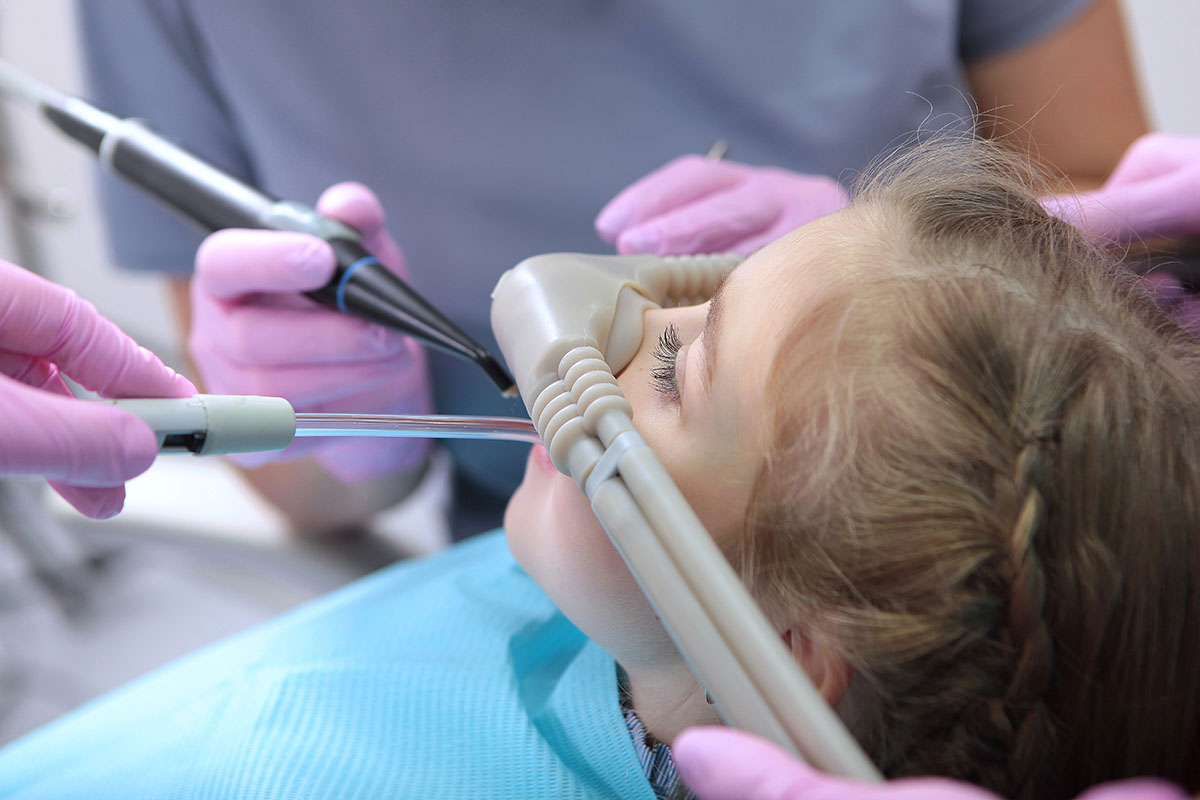 A young patient is lying in a dental chair with a dental professional using tools, while another staff member assists. The patient is wearing a protective bib and appears calm during the procedure.