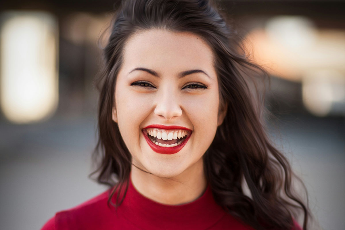 A woman with long, wavy dark hair smiles broadly, showcasing bright red lipstick and a joyful expression while wearing a high-neck, red top. The background is softly blurred, emphasizing her face.