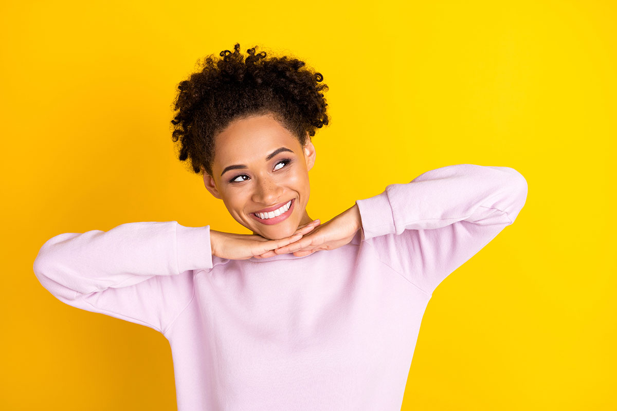 A smiling young Black woman wearing a light pink sweatshirt resting her chin on her hands against a bright yellow background.