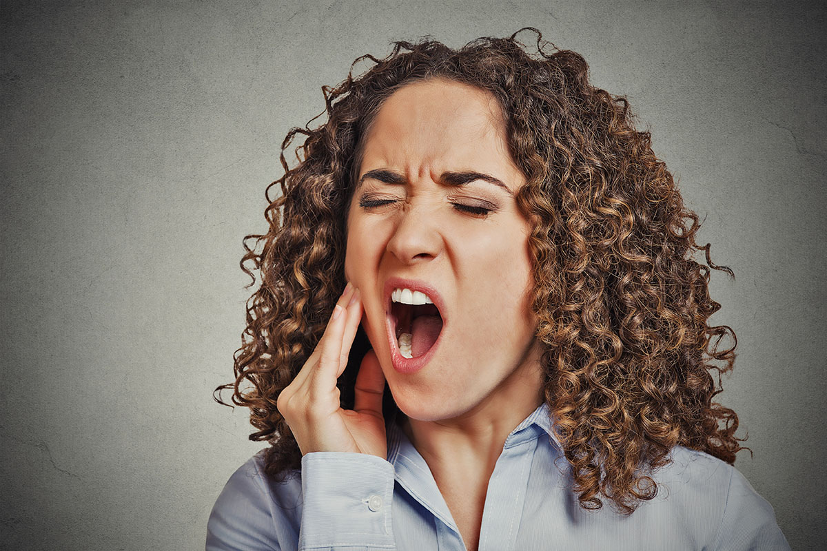 A woman with curly hair is grimacing and holding her jaw in pain, appearing to yawn. She wears a light blue shirt, and the background is a textured gray.