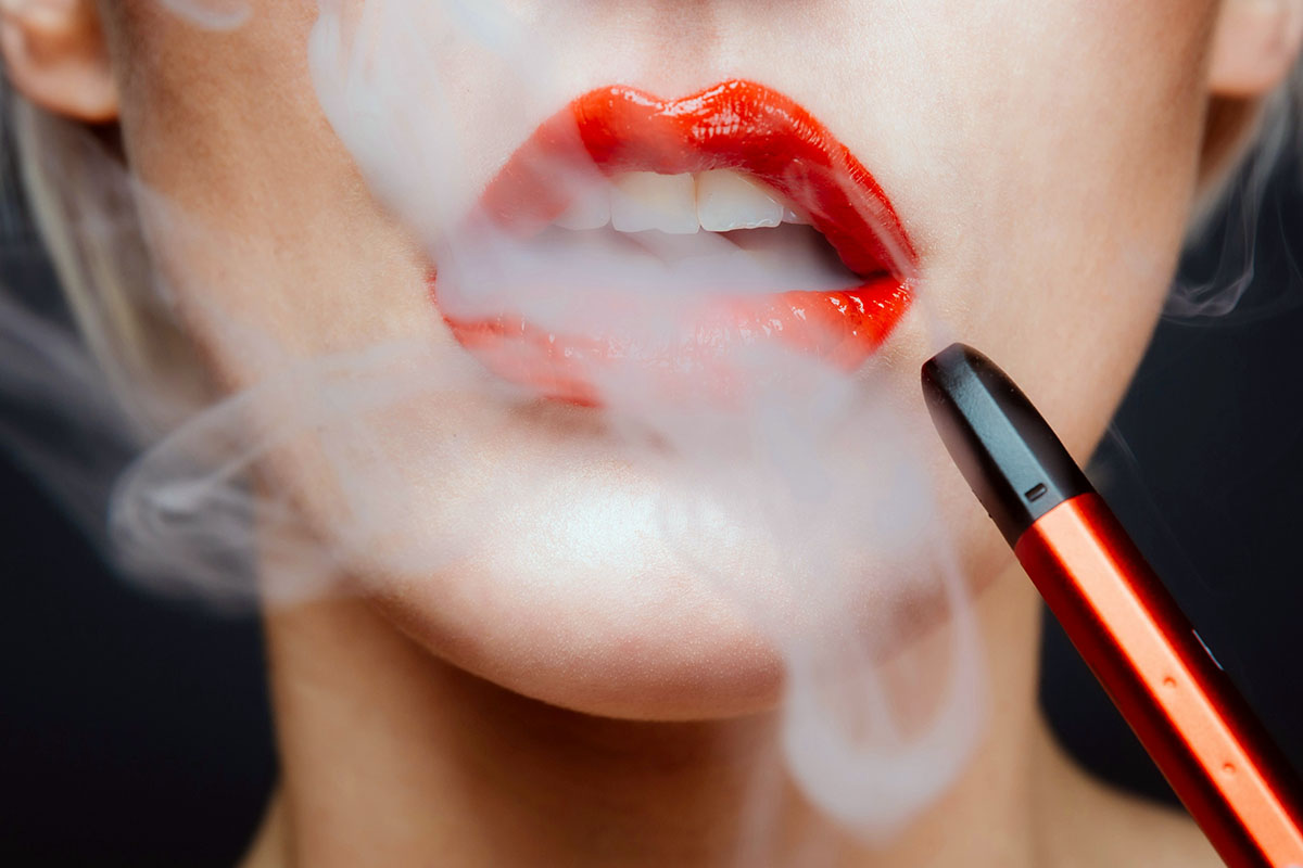 Close-up of a person with red lipstick exhaling a cloud of vapor while holding an orange electronic vape pen.