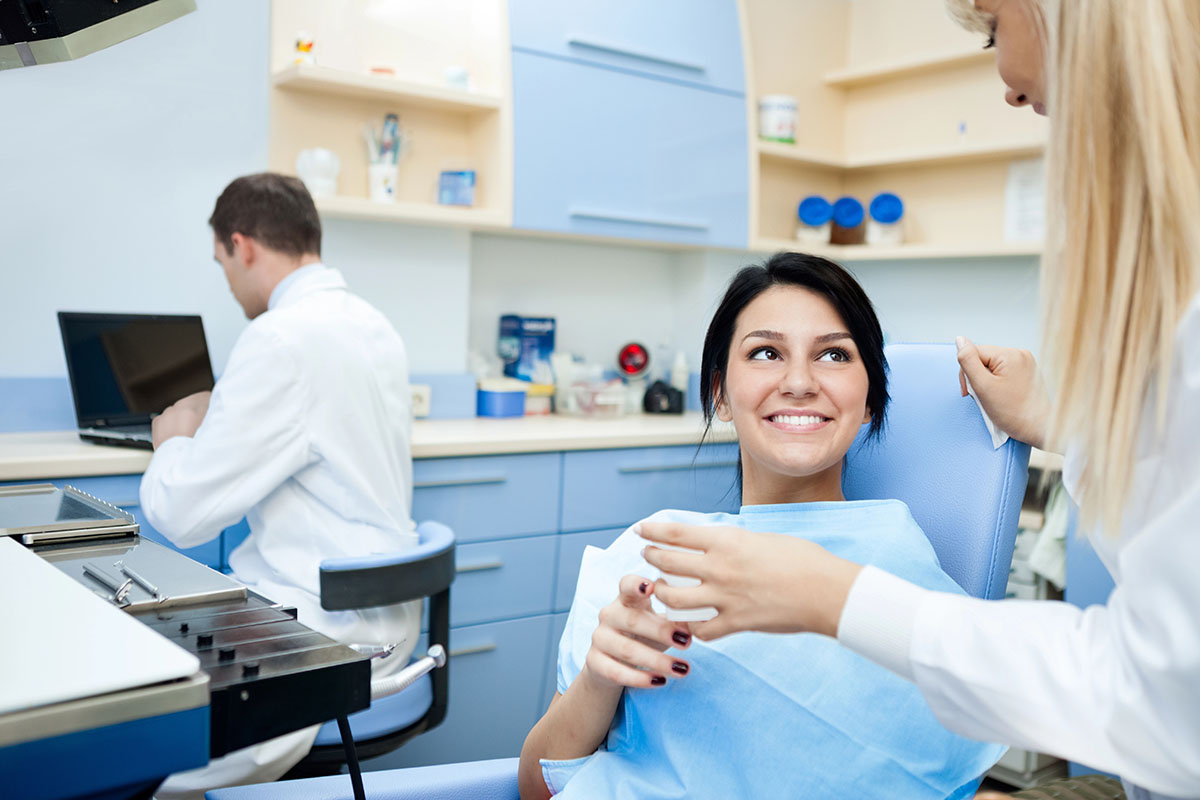 A woman sitting in a dental chair, smiling and wearing a blue dental bib, while a dental professional speaks to her. In the background, a male dentist is using a laptop. The dental office has light blue walls and clinical equipment visible.
